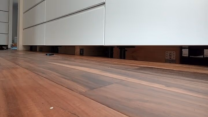 Nhìn sát mặt sàn gỗ vào chân một dãy tủ trắng chưa lắp len chân: dưới đáy tủ là các chân nhựa đen đứng thành hàng, phía sau lộ ra chân tường và vài tấm gỗ.