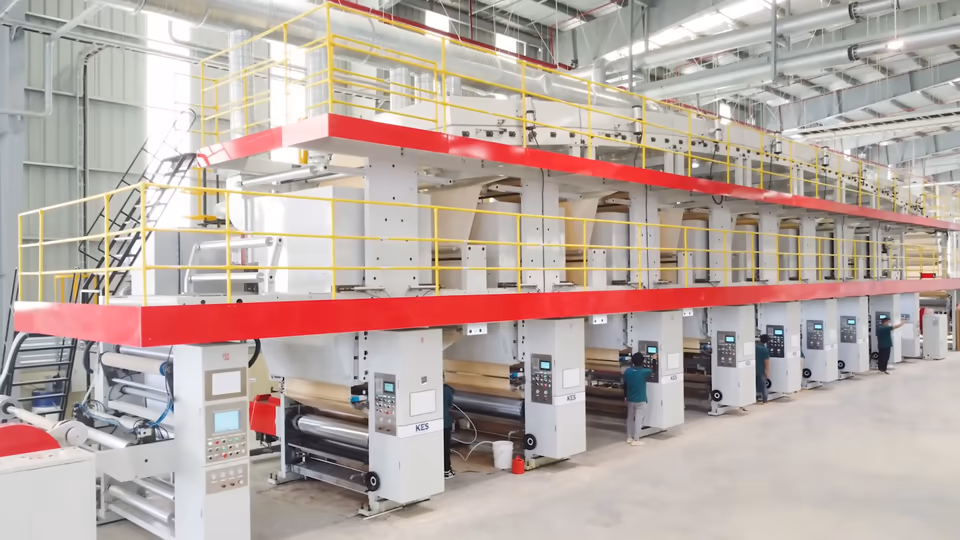 Dãy máy in nhiều cụm xếp dài trong nhà xưởng, hai tầng lan can vàng và dải đỏ. Ở tầng dưới, công nhân mặc áo xanh đứng cạnh các bảng điều khiển có chữ KES, giấy nền màu be chạy qua các trục.