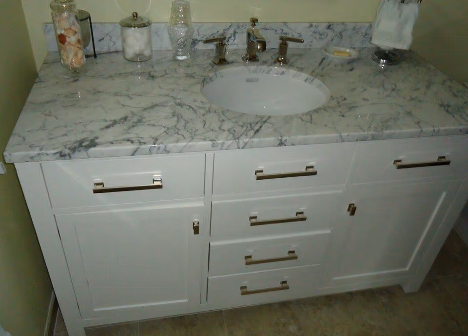 Một tủ lavabo đặt sàn sơn trắng, mặt đá vân trắng xám có chậu âm và một tấm đá ốp chắn sát tường phía sau. Bốn trụ góc của tủ đứng thẳng xuống sàn gạch, thanh ngang dưới cùng nằm sát mặt sàn.