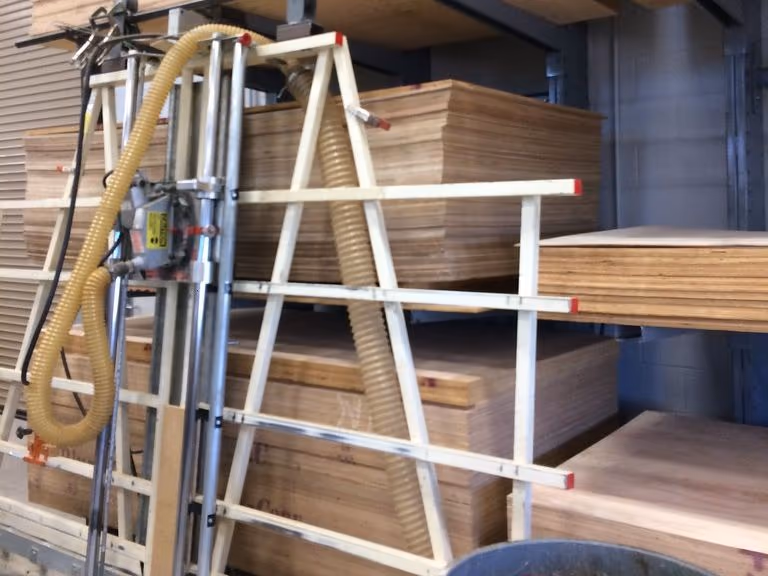Một máy cưa panel đứng khung sơn trắng có ống hút bụi màu vàng, đặt trước giá để ván. Trên giá là nhiều chồng ván plywood nguyên khổ xếp nằm, nhìn rõ cạnh các tấm chồng lên nhau.
