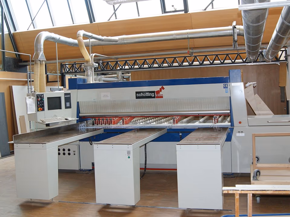 Máy cưa panel nằm ngang trong một xưởng trường nghề: thân máy dài có thanh ép phía trên, phía trước là ba bàn đỡ ván, bên trái có màn hình điều khiển. Dọc tường bên trái dựng vài tấm ván nguyên khổ.