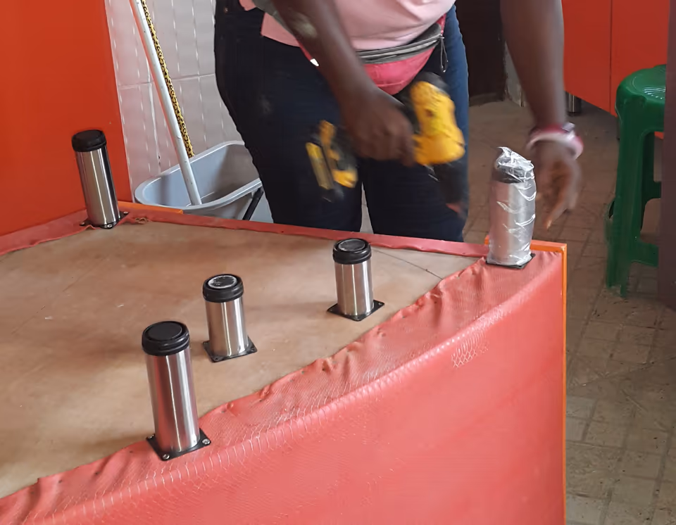 Một thùng tủ bếp đặt lật ngửa, đáy tủ quay lên trên. Năm chân tủ hình trụ bằng inox đã bắt vào đáy, mỗi chân có đế vuông bắt vít. Phía sau, người thợ cầm máy khoan bắt vít, tay kia giữ chân tủ cuối cùng.