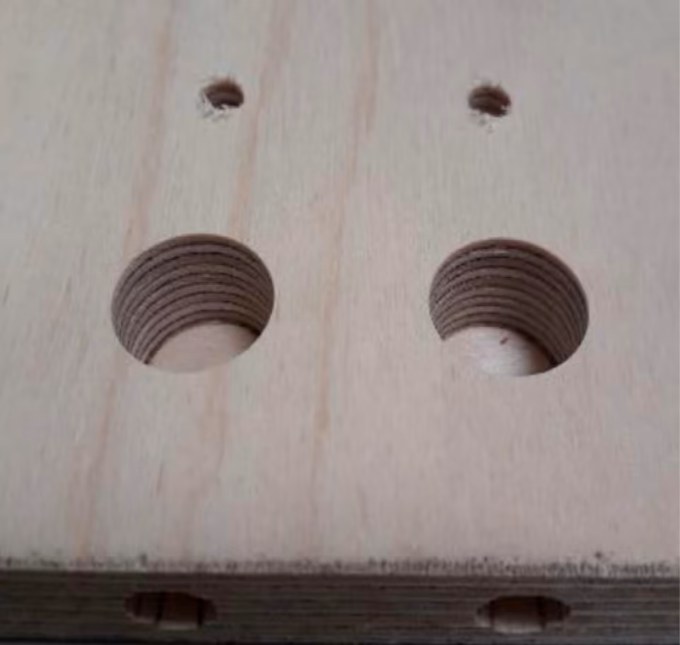 Mặt một tấm ván plywood bạch dương sáng màu có hai lỗ tròn lớn khoan không thủng, thành lỗ lộ các lớp veneer xếp chồng; ở cạnh tấm phía dưới có hai lỗ nhỏ khoan dọc vào, thẳng hàng với hai lỗ lớn.
