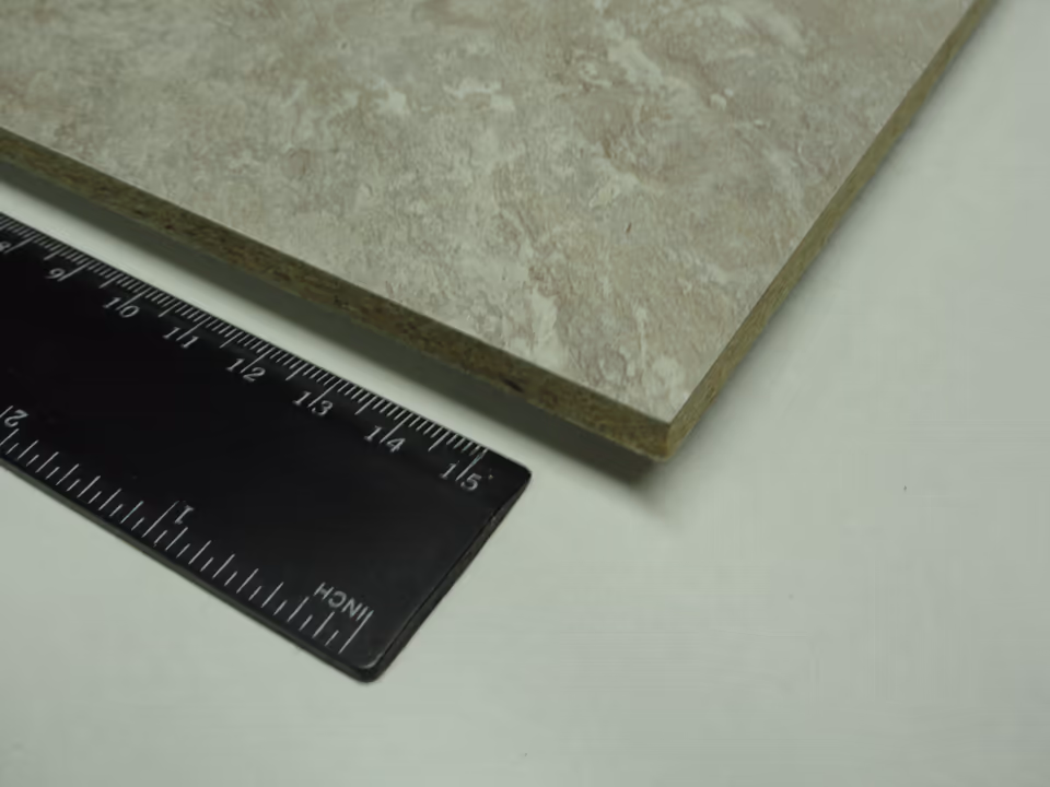 Góc một tấm ván phủ laminate vân đá màu be đặt cạnh thước kẻ. Mép cắt lộ lớp cốt ván dăm màu nâu xám, trên cùng là đường mỏng của tấm laminate.