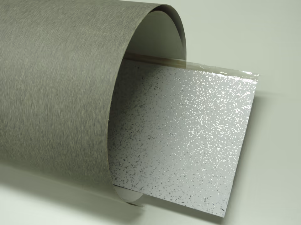 Một tấm laminate màu xám vân xước mỏng đến mức cuộn tròn được như tờ giấy cứng, bên trong lòng cuộn lộ một tấm laminate khác màu trắng có hạt ánh bạc.