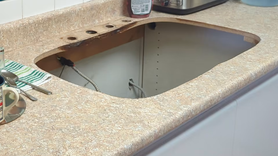 Một mặt bàn bếp phủ laminate vân đá đã tháo chậu rửa, để lộ lỗ khoét có góc bo tròn và mép cắt lộ phần cốt ván dăm màu nâu chưa được bịt kín.