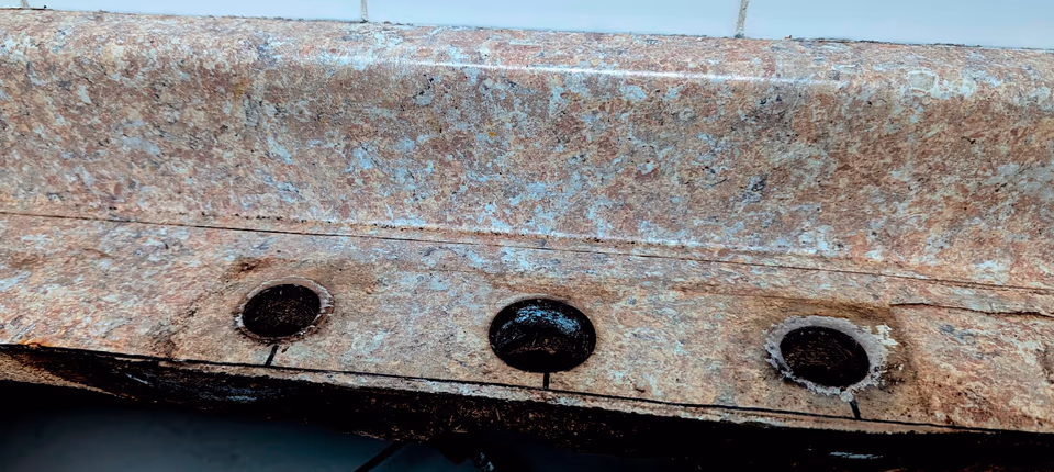 Mép sau lỗ khoét chậu rửa trên một mặt bàn bếp phủ laminate vân đá, chậu đã tháo ra: ba lỗ lắp vòi có lõi ván sẫm đen, mặt laminate nứt thành đường dọc mép và tỏa ra từ các lỗ, cốt ván dăm ở mép cắt sẫm màu và bở ra.
