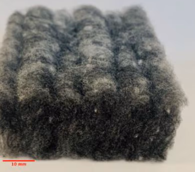Một khối sợi polyester màu xám đen chụp ngang ở cự ly gần, các sợi mảnh rối vào nhau, nhìn rõ là vật liệu rất xốp; góc dưới có vạch tỷ lệ 10 mm.